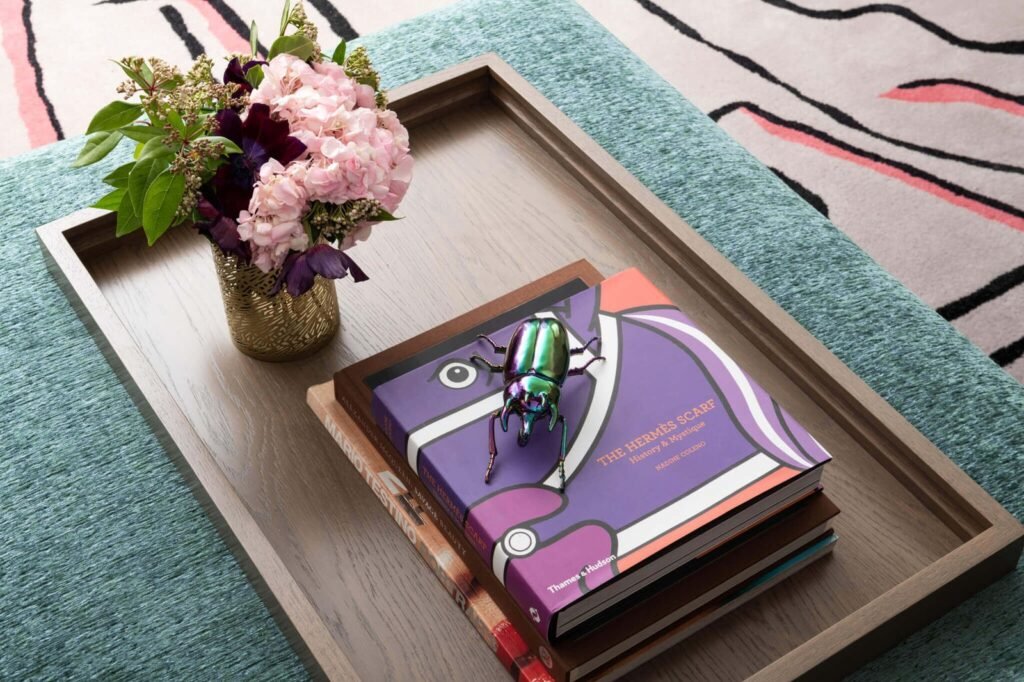 Stylish coffee table tray with fashion books, iridescent beetle figurine, and a gold vase of pink & purple flowers.