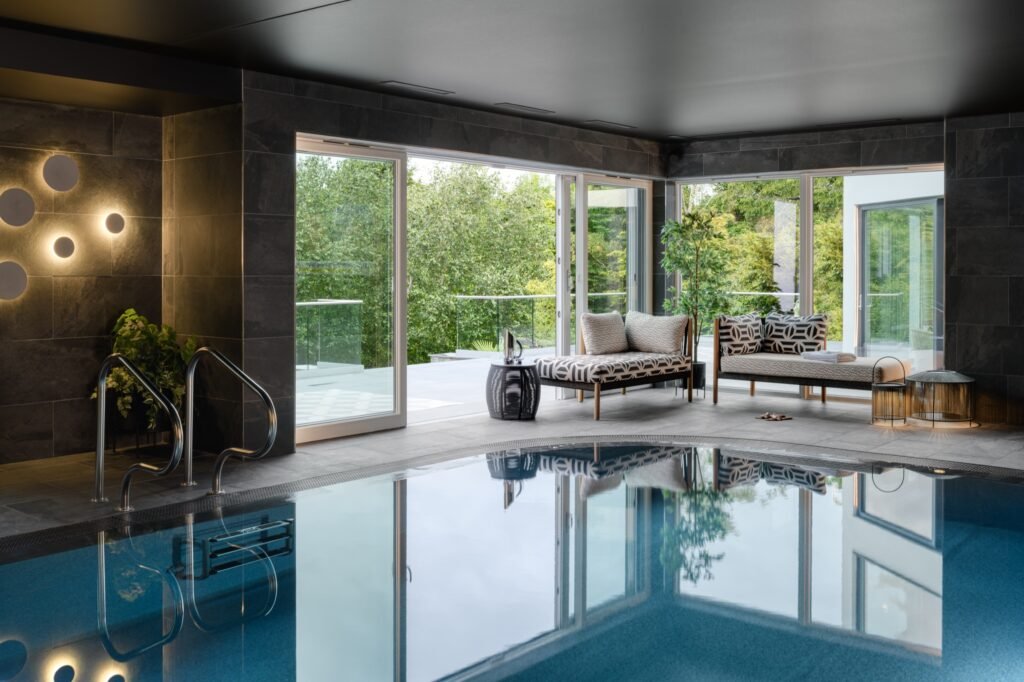 Luxurious indoor pool with dark textured walls, stylish lounge area, and glass doors opening to a lush forest view.