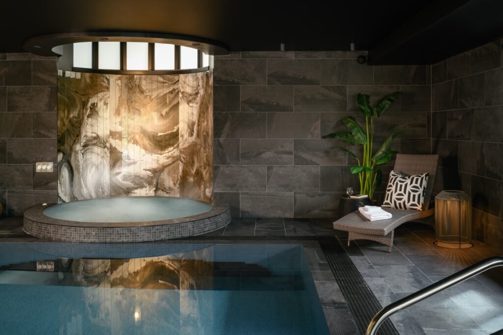 Modern indoor pool and hot tub with dark slate tiles, a curved marble accent wall, and a lounge chair.