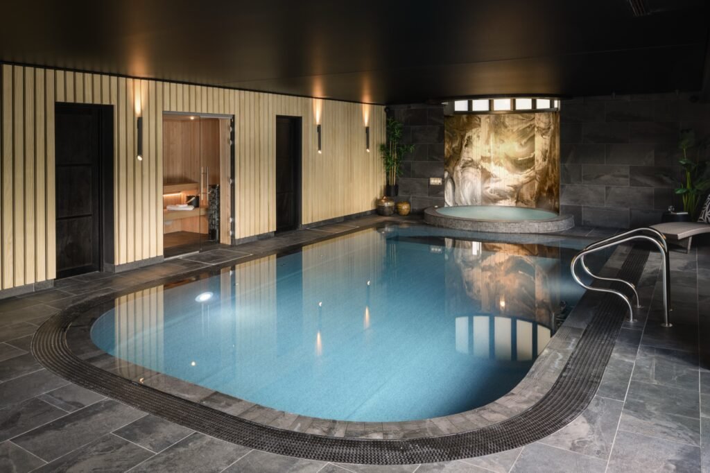 Elegant indoor swimming pool and jacuzzi spa area with a sauna, light wood paneling, dark stone tiles, and ambient lighting.