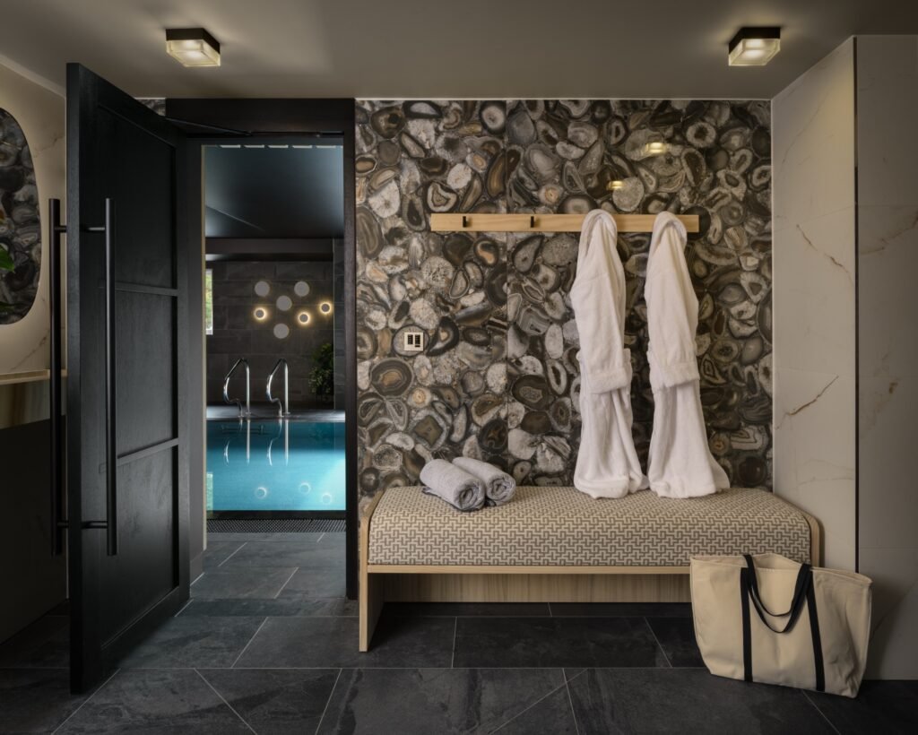 Stylish spa changing area with agate wall, patterned bench, bathrobes, and view of indoor pool through open door.
