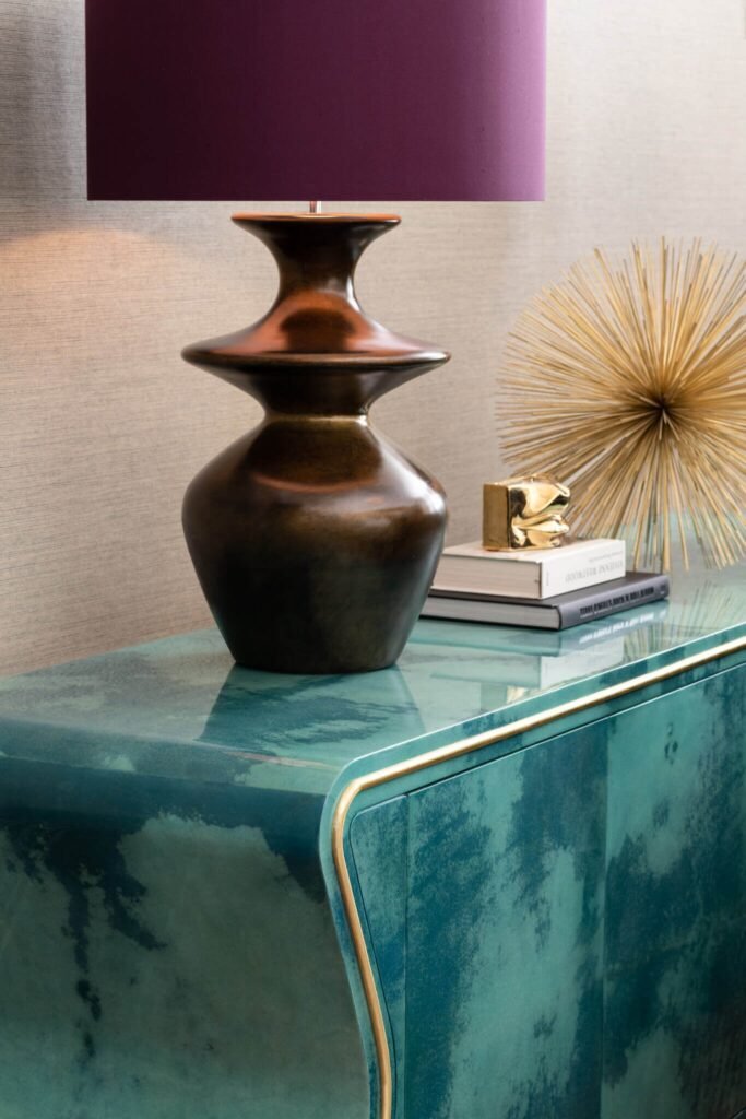 Teal-green console with gold trim, bronze lamp, purple shade, and modern gold decor on textured wallpaper.