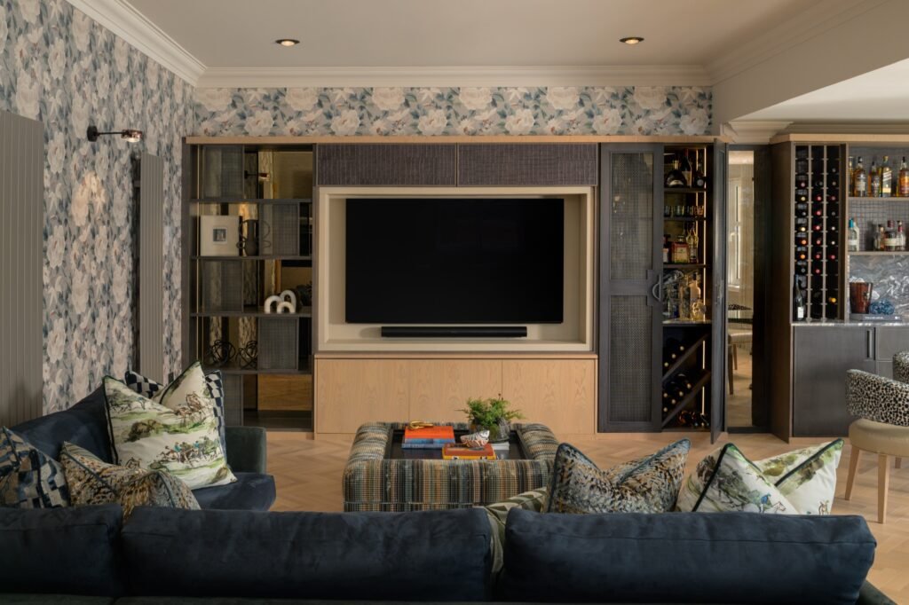 Elegant living room with floral wallpaper, custom TV unit, integrated bar, navy sofa, plaid ottoman, and herringbone floor.