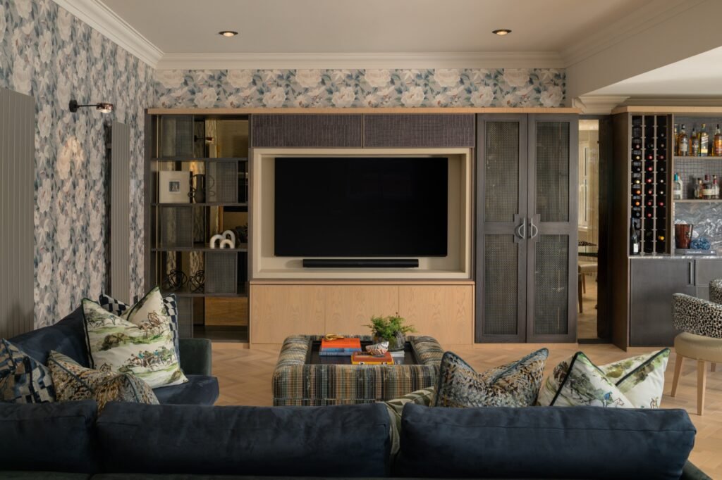Elegant living room with floral wallpaper, built-in media unit, dark blue velvet sofa, plaid ottoman, and wet bar.