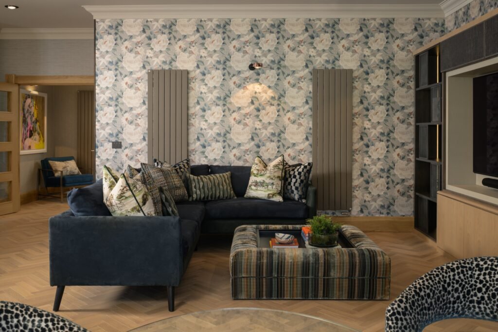 Elegant living room with white and blue floral wallpaper, dark blue velvet sofa, plaid ottoman coffee table, and herringbone wood floor.