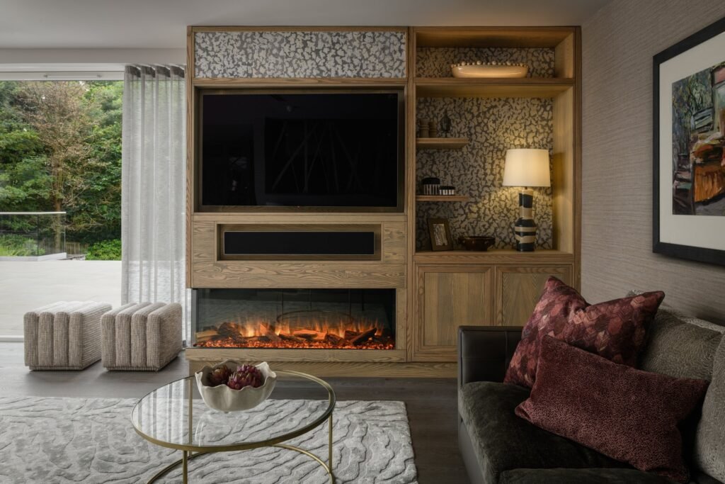 Elegant living room with built-in wooden media unit, TV, glowing electric fireplace, dark sofa, glass table, and nature view.