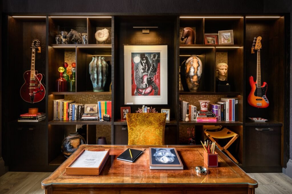 Richly detailed home office with a classic wooden desk, dark wood shelves filled with books, eclectic art, and guitars.