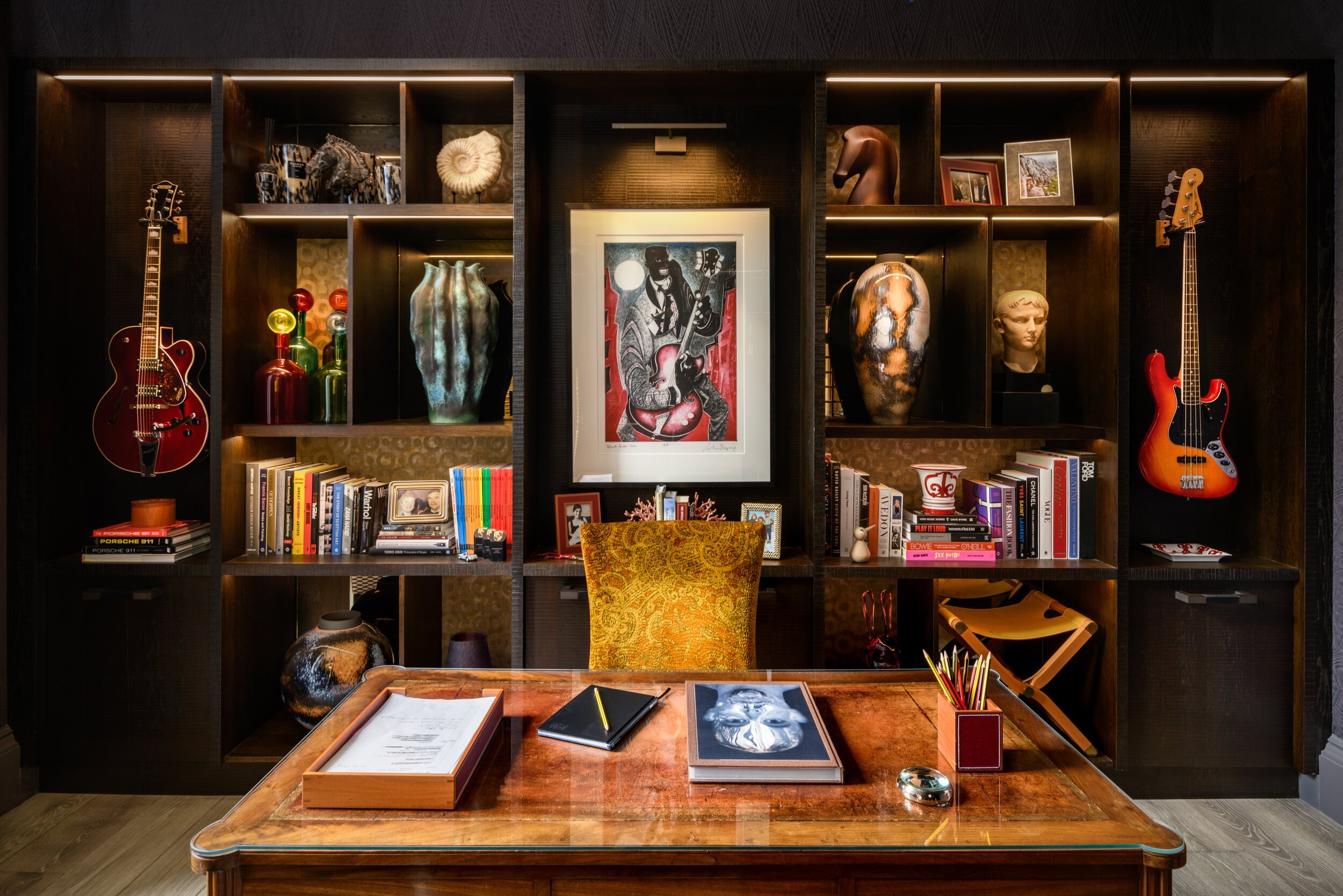 Richly detailed home office with a classic wooden desk, dark wood shelves filled with books, eclectic art, and guitars.