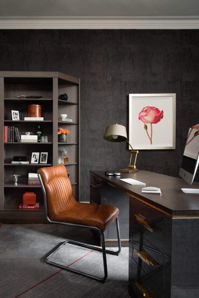Modern home office with dark textured walls, a dark wood desk, brown leather chair, and a bookcase. Rose art hangs above.