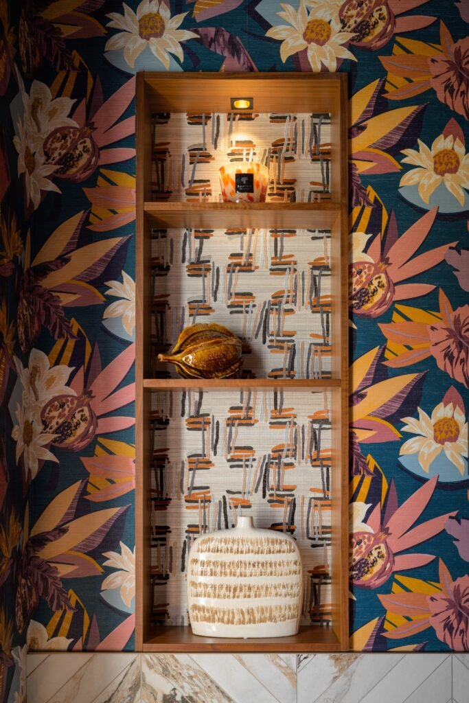 Vibrant tropical wallpaper flanks a wooden niche with an abstract-patterned back, displaying decorative objects and accent lighting.