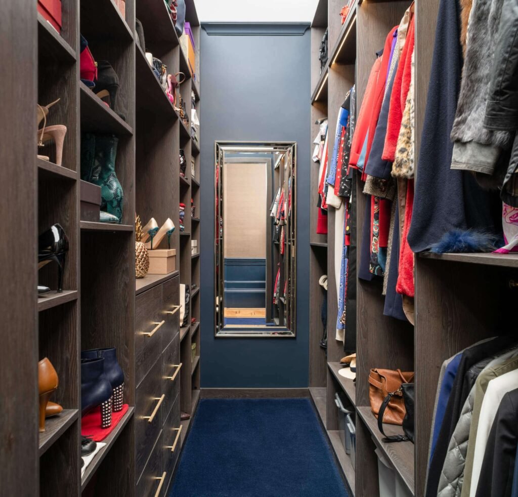 Elegant dark wood walk-in closet with shelves of shoes & accessories, hanging clothes, a full-length mirror, and navy rug.