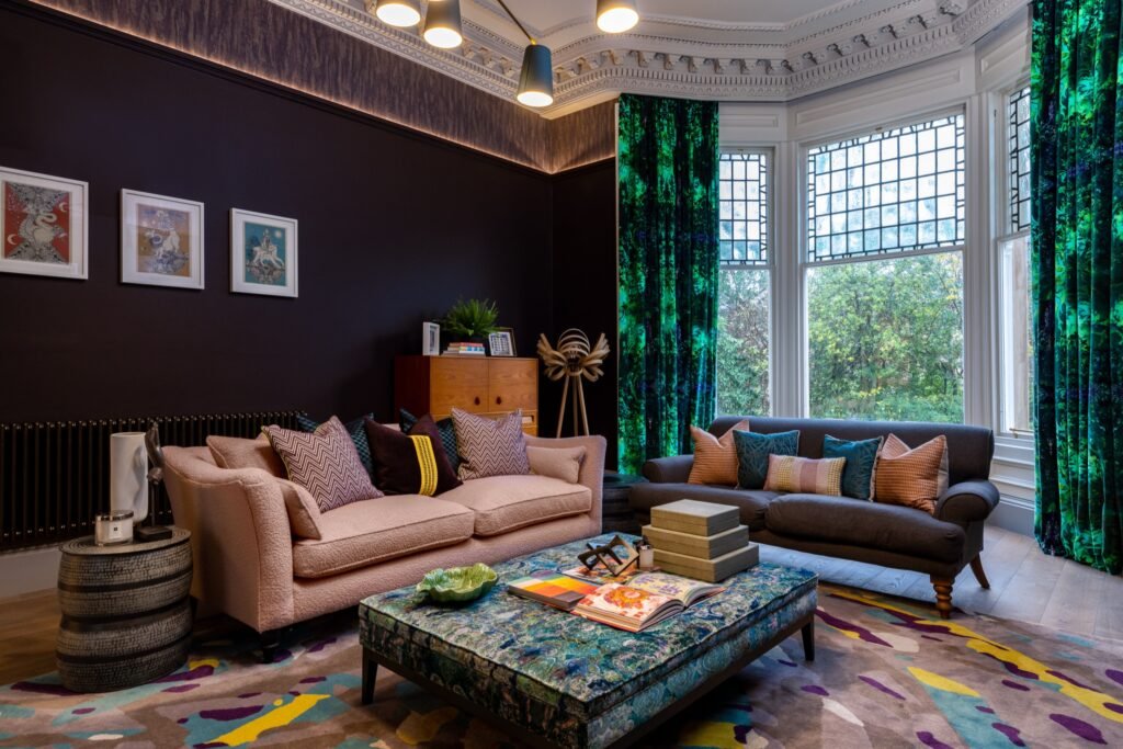 Eclectic living room with dark walls, vibrant green curtains, colorful rug, pink & gray sofas, ornate ceiling.