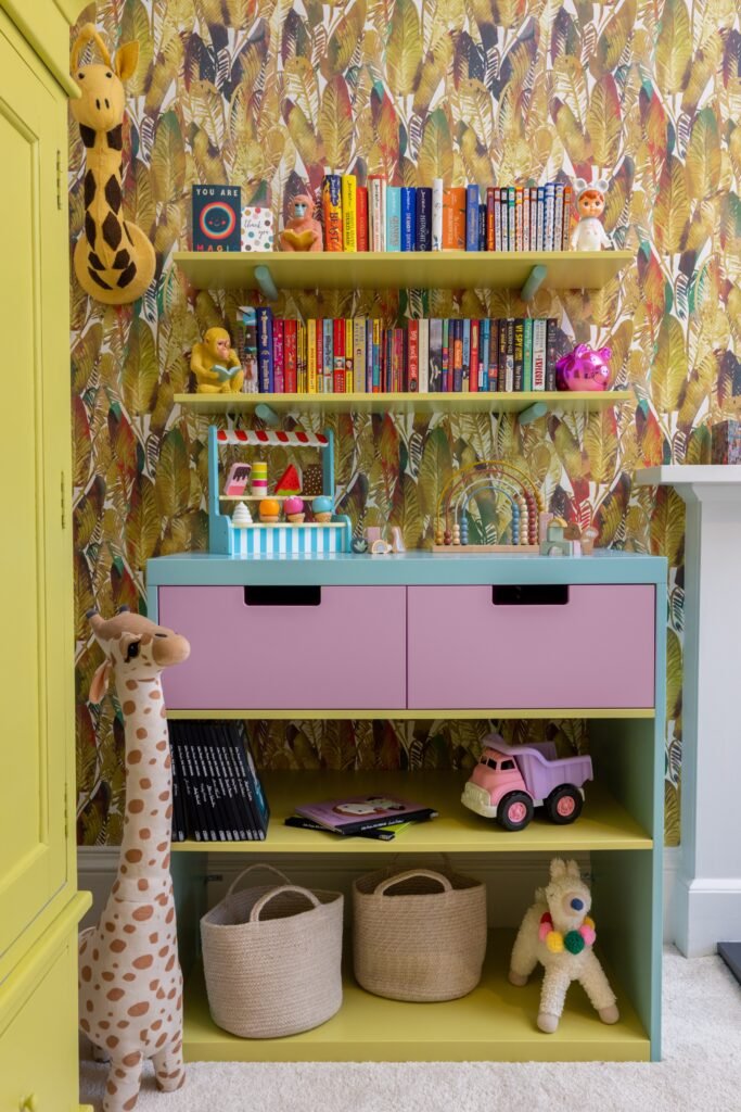 Vibrant children's room with jungle leaf wallpaper, yellow shelves filled with books and toys, and a pastel blue and pink cabinet.