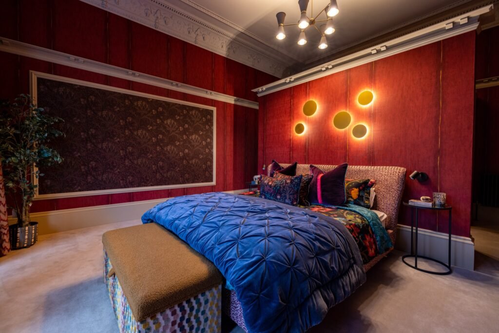 Bold bedroom with rich red walls, gold sconces, a blue velvet bed, floral bedding, and a decorative patterned bench.
