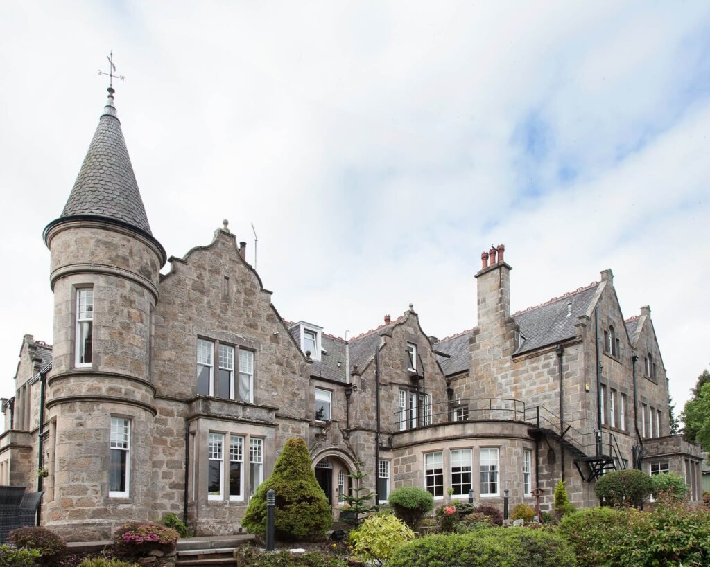 Exterior of a historic stone building with a prominent round tower, conical roof, and landscaped entrance.