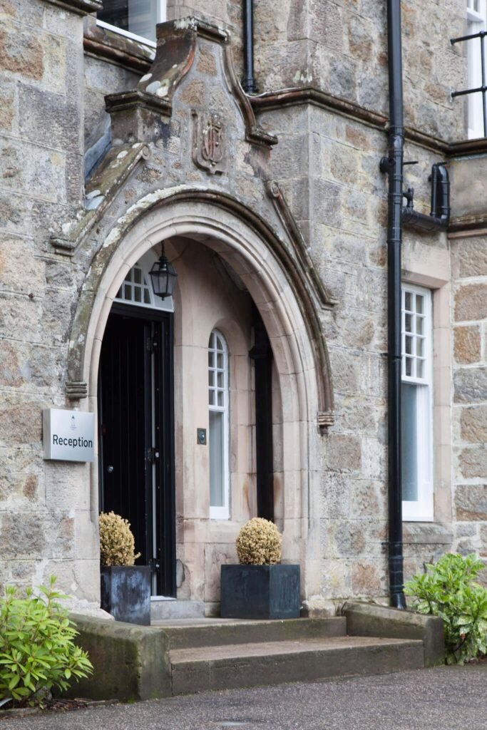 Weathered stone building entrance with an ornate arched doorway, crest, black door, 'Reception' sign, and planters.