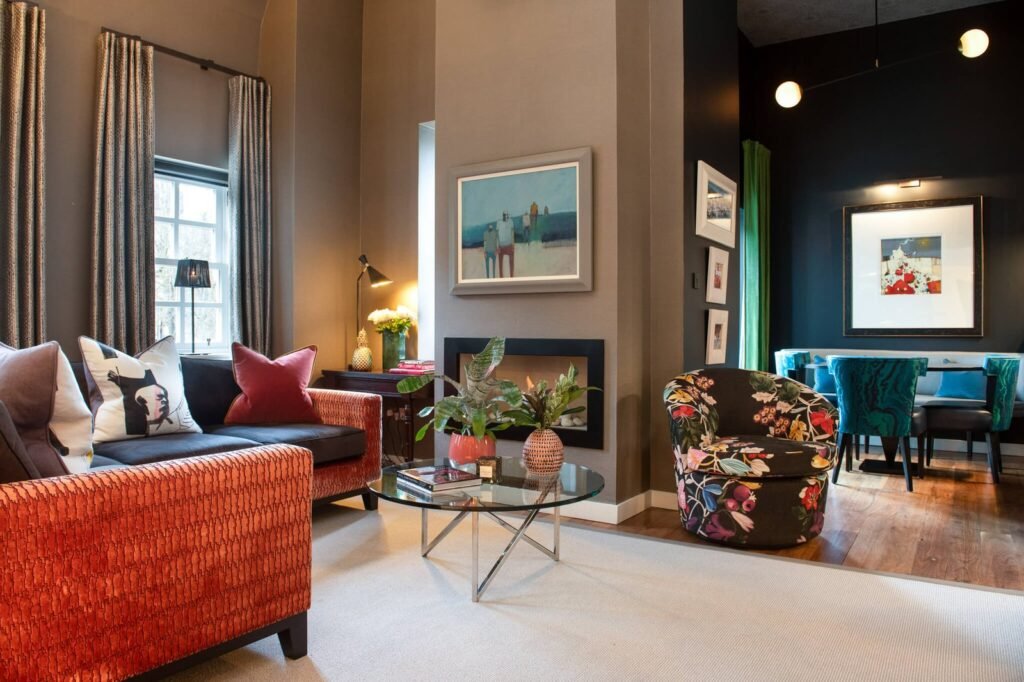 Vibrant, eclectic living and dining space with orange sofas, floral armchair, modern fireplace, and colorful artwork.