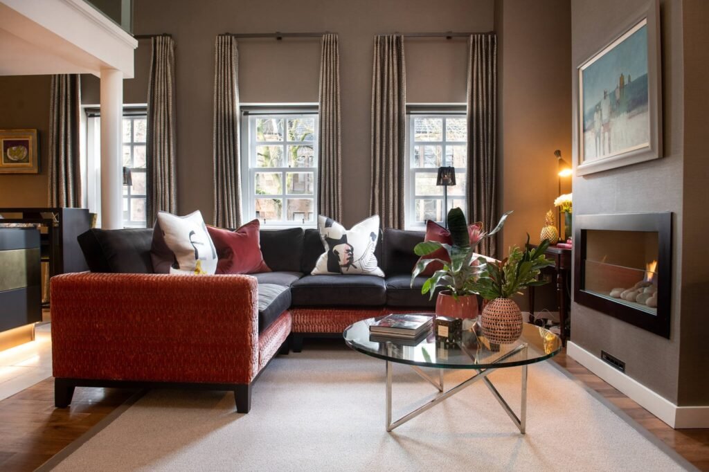 Warm modern living room featuring a dark L-shaped sofa with red accents, glass coffee table, contemporary fireplace, and plants.