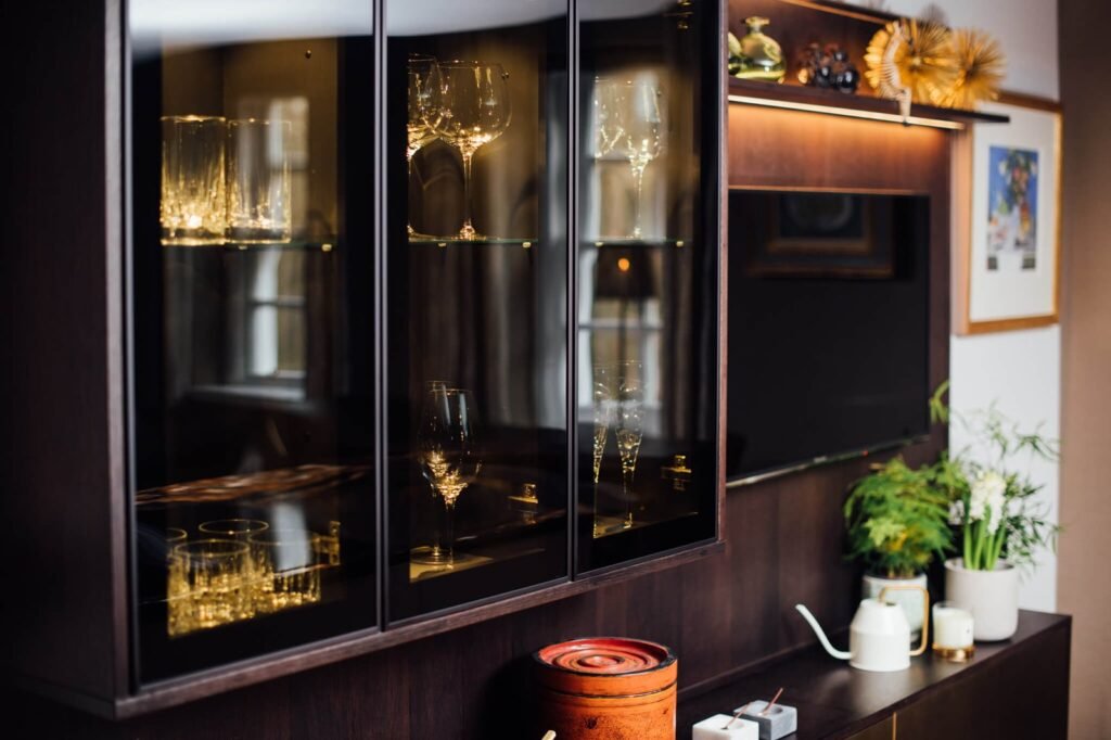 Elegant dark wood display cabinet showcasing illuminated crystal glassware, with a TV, plants, and decor.