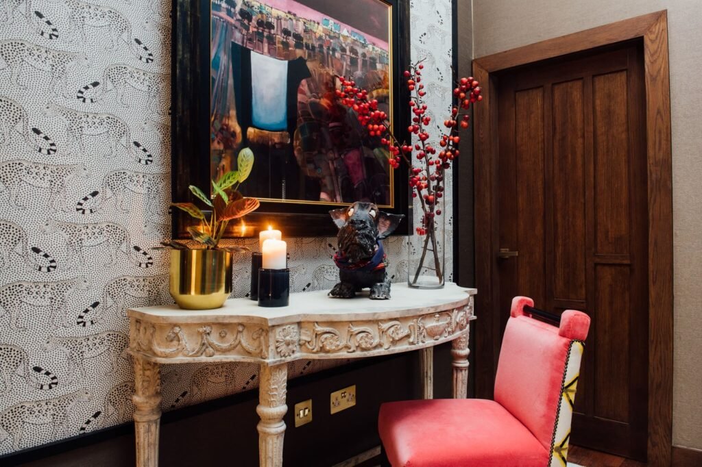 Stylish eclectic hallway with ornate console, cheetah wallpaper, abstract art, dog statue, red berries, and a pink chair.