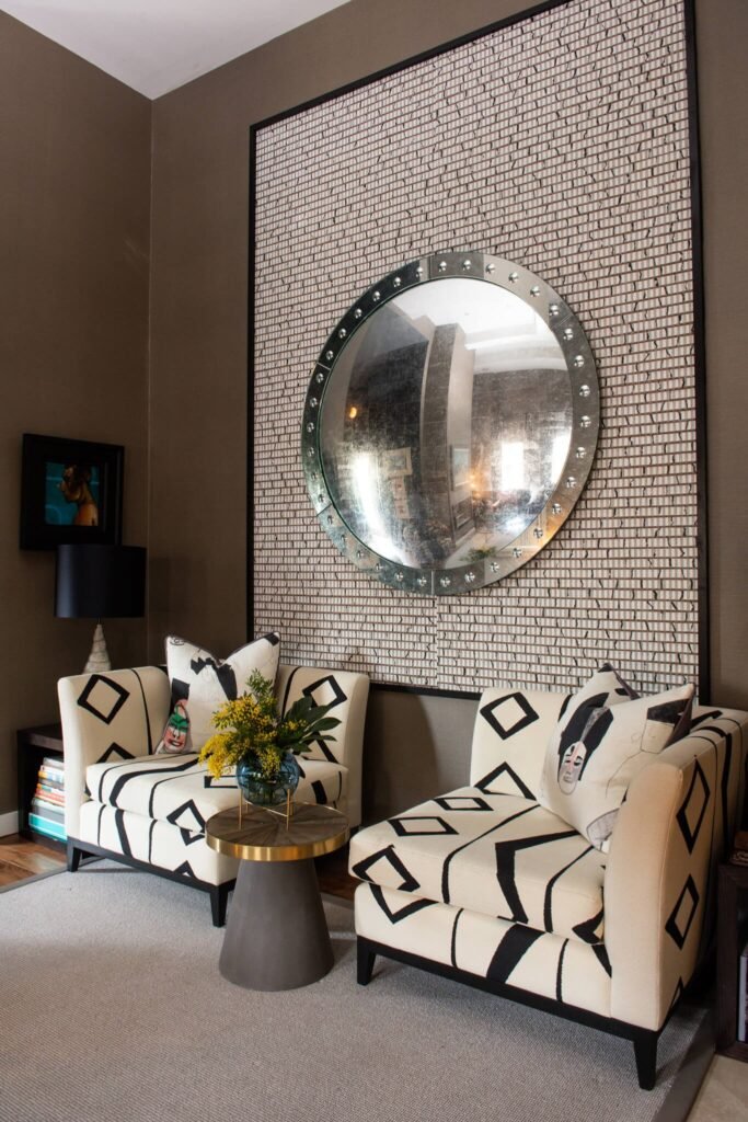 Modern eclectic seating area with geometric armchairs, a large studded porthole mirror over a textured panel, and a conical side table.
