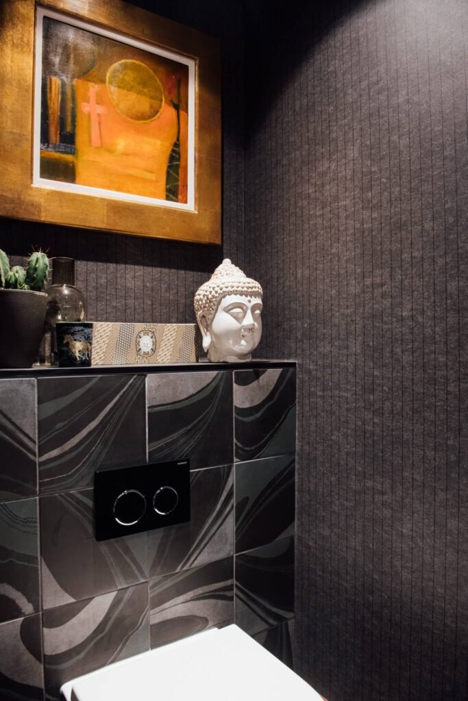 Modern powder room with dark striped wallpaper, swirled black tiles, a Buddha head, and abstract art with a gold frame.