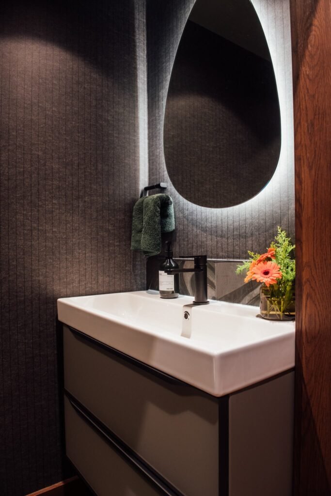 Modern powder room with dark textured wallpaper, white sink, matte black faucet, and backlit organic mirror.