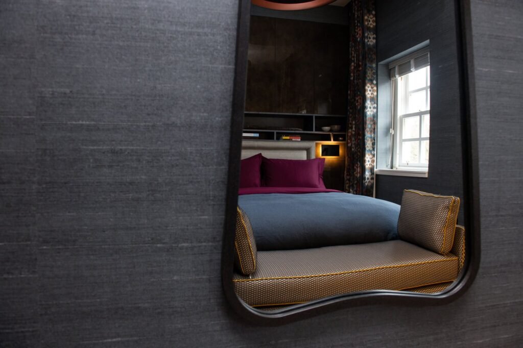 Moody bedroom interior reflected in a mirror, showing a bed with plum pillows, blue duvet, built-in shelves, and a patterned daybed.