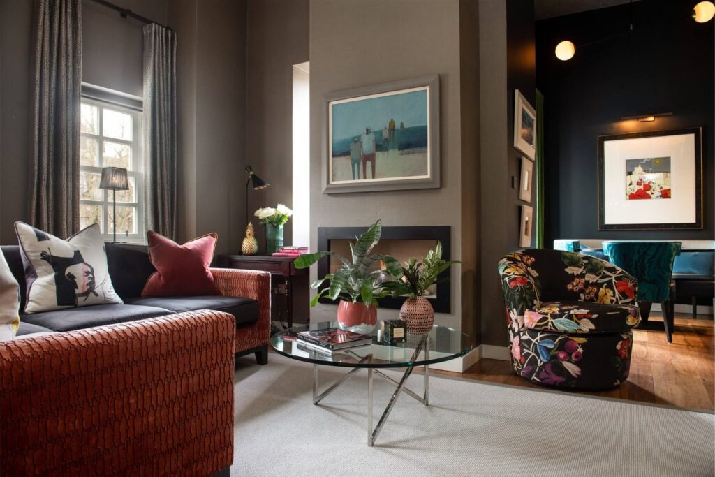 Eclectic living room with dark walls, orange sofa, bold floral armchair, fireplace, glass coffee table, and vibrant art.