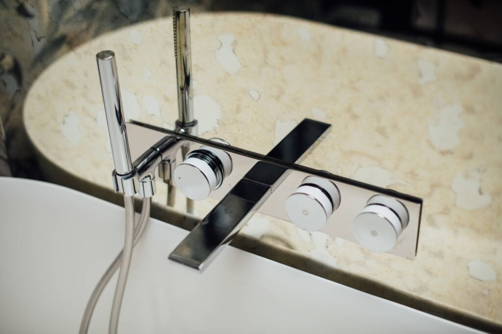 Sleek chrome bathtub filler with wide spout, white/chrome control dials, and handheld shower on light marble.