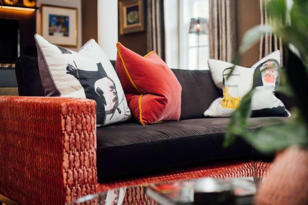 Dark sofa with vibrant red velvet and artistic patterned cushions, next to a textured orange armrest in a stylish living room.