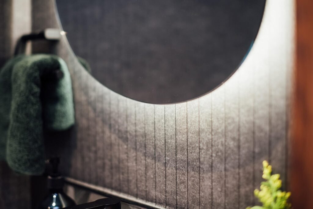 Modern bathroom detail: a round backlit mirror illuminates a dark textured wall, with a green towel and black fixtures.