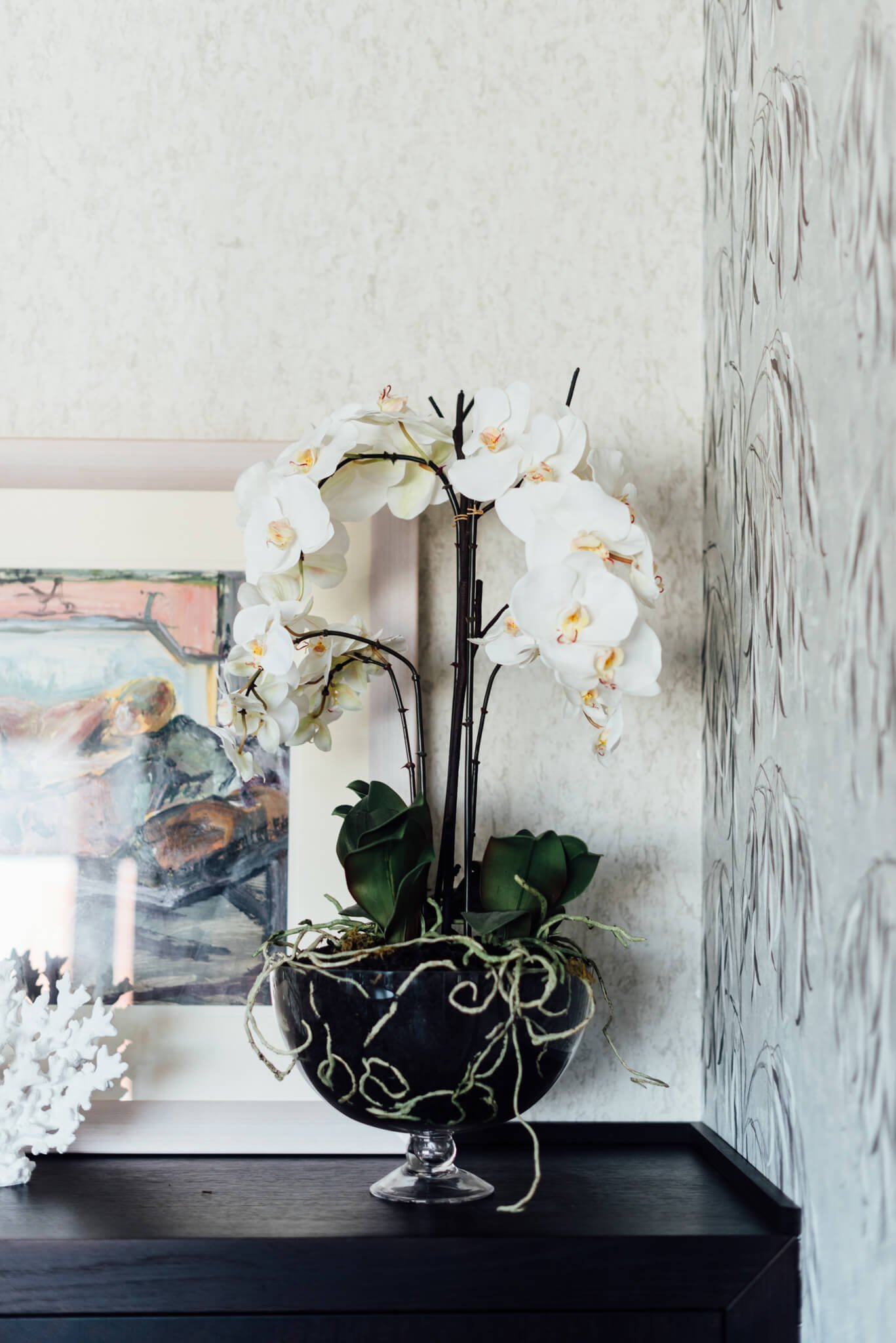 Elegant white orchid in a dark planter on a black console table, with textured wallpaper and abstract art.