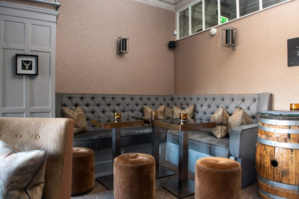 Elegant lounge bar with grey tufted banquette, sleek square tables, velvet stools, geometric wallpaper, and a whiskey barrel.