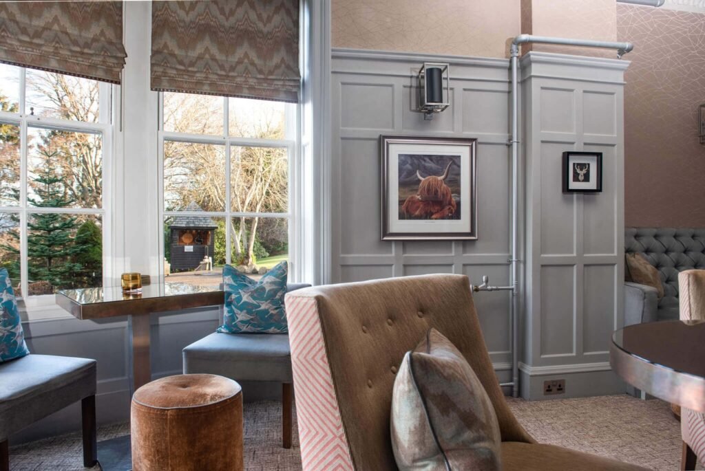 Cozy grey paneled lounge with large windows, Highland cow art, stylish armchairs, and a garden view.