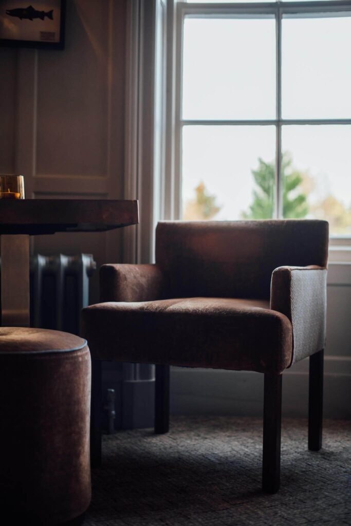 Cozy lodge interior with a plush brown armchair, matching ottoman, and table with a drink by a window with tree views.