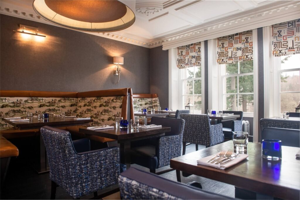 Modern classic restaurant with dark textured walls, scenic toile booths, blue armchairs, wood tables, geometric blinds.