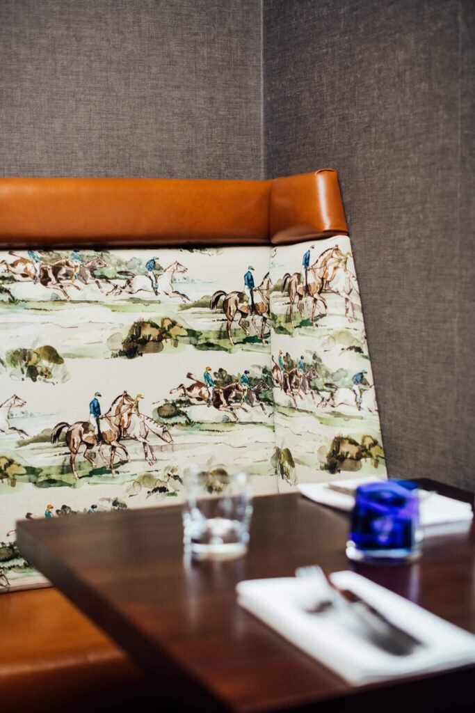 Brown leather and horse racing print booth in a restaurant. Dark wood table set with a glass, blue tumbler, and cutlery.