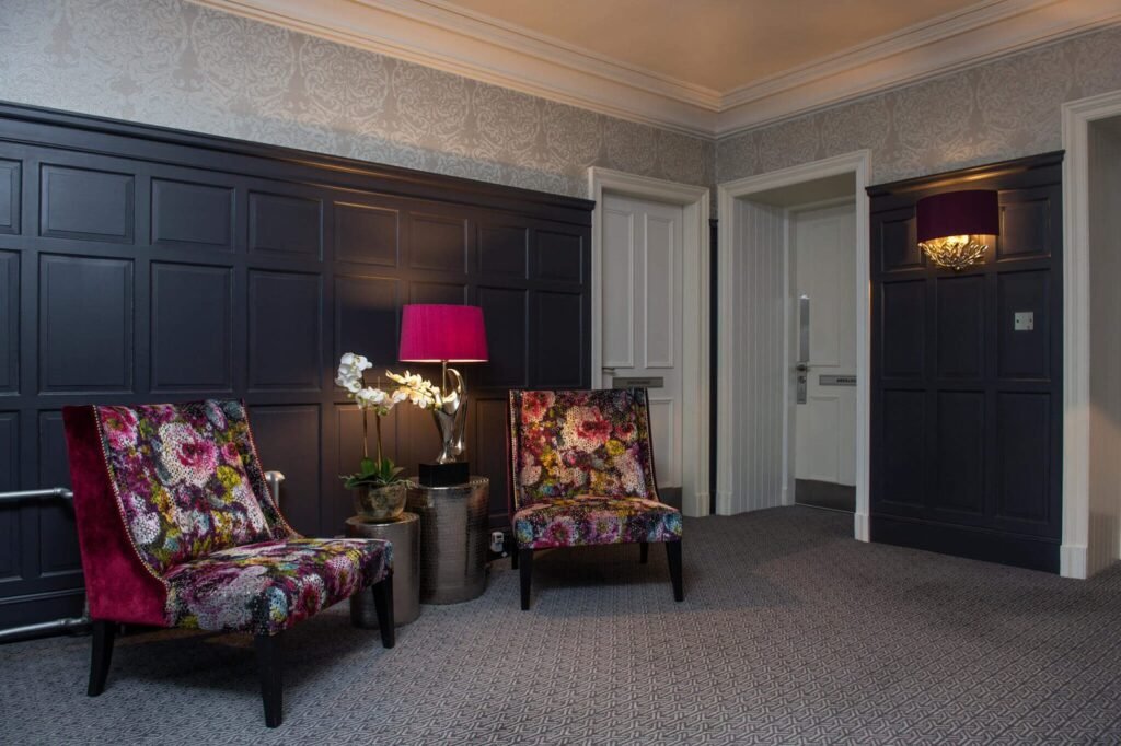 Stylish lounge with dark paneling, patterned wallpaper, two vibrant floral chairs, bright pink lamp, and gray carpet.