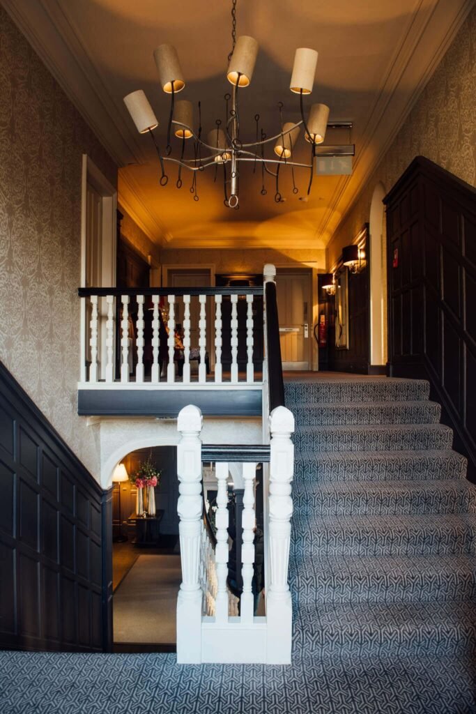 Warmly lit grand hall with dark wood paneling, white balusters, patterned gray carpeted stairs, and elegant chandelier.