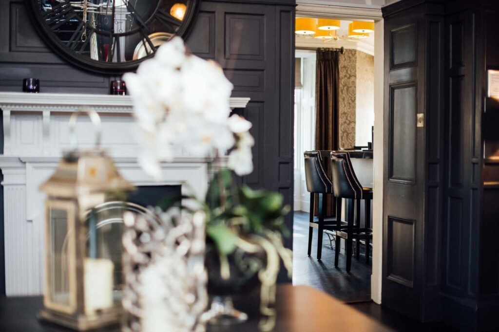Sophisticated dark paneled lounge with fireplace, mirror, leading to a stylish bar area with black studded stools and yellow lights.