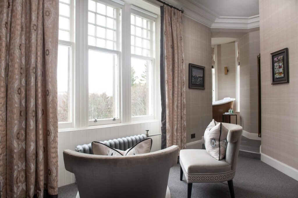 Elegant suite with large window, patterned curtains, two armchairs, and freestanding bronze tub visible beyond.