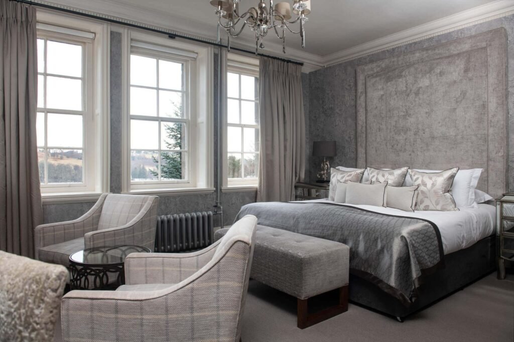 Luxurious grey bedroom with a large bed, upholstered headboard, ornate chandelier, two armchairs, and scenic windows.