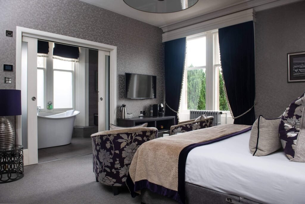 Elegant bedroom with a large bed, purple floral armchairs, TV, and a freestanding bathtub visible through sliding doors.