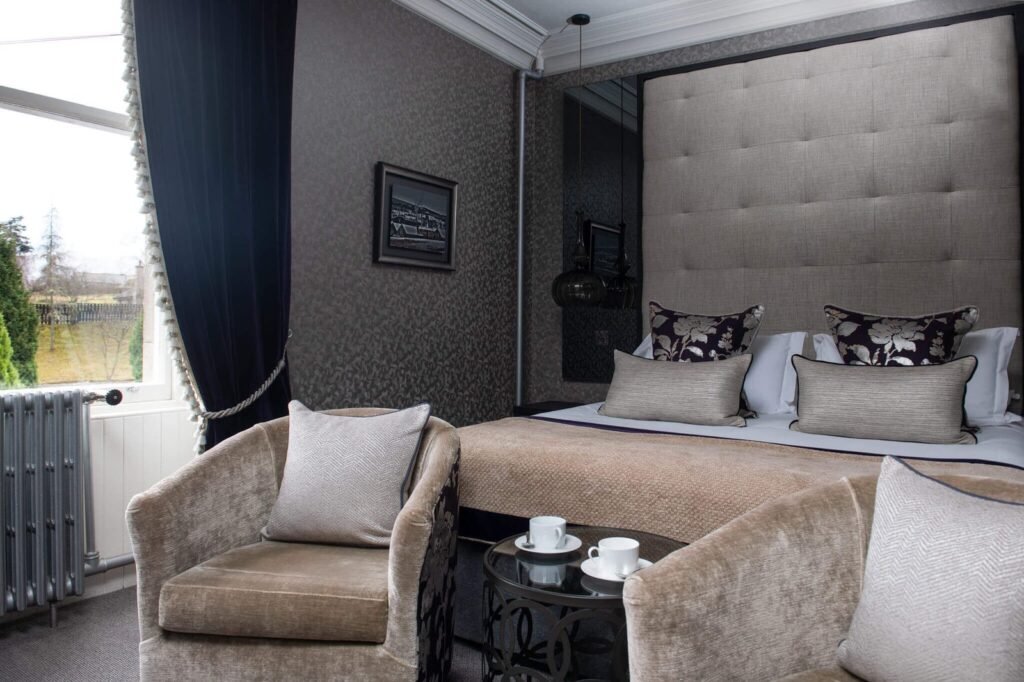 Elegant hotel bedroom featuring a beige tufted bed, two velvet armchairs with coffee, dark patterned walls, and blue curtains.