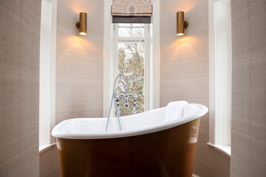 Elegant brown and white freestanding bathtub in a bright bathroom alcove under a window, with warm brass sconces and textured walls.