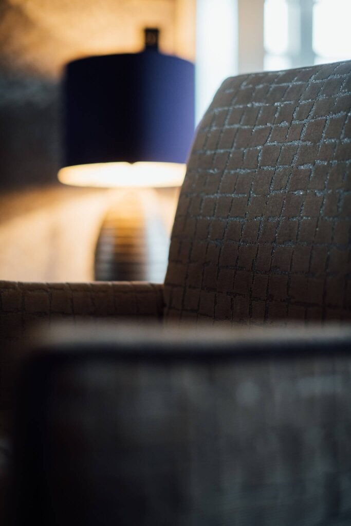 Close-up of dark geometric patterned armchair. Blurry blue lampshade illuminates a ribbed lamp base with warm light.