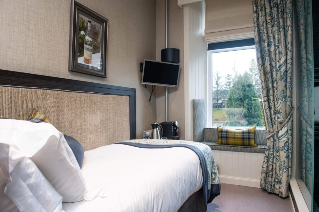 Cozy single bedroom with textured headboard, tartan window seat cushion, floral curtains, and a scenic view.