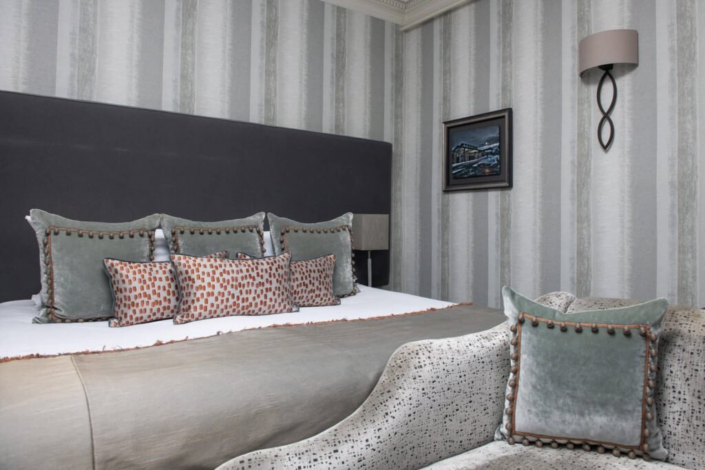 Elegant bedroom with striped wallpaper, dark headboard, velvet pillows, and patterned bench. Neutral tones with orange accents.