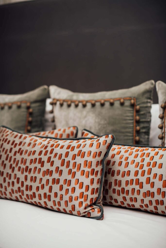 Close-up of modern decorative pillows: orange brushstroke pattern with piping, and grey velvet with pom-pom trim.